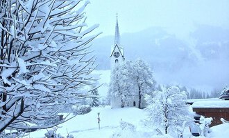 Eine verschneite Landschaft mit einer Kirche im Hintergrund. Bäume und Sträucher sind vollständig mit Schnee bedeckt.
