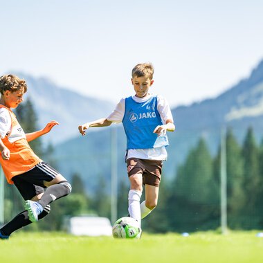 Junge Fußballspieler auf einem grünen Spielfeld mit Bergen im Hintergrund. Ein Spieler dribbelt den Ball, während ein anderer ihn versucht zu stoppen. | © Fußballschule Anstoß