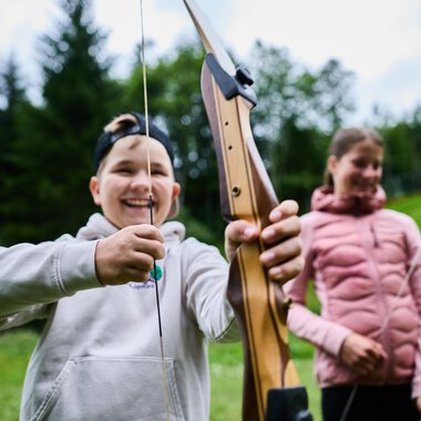 Two people in the woods shooting arrows. The woman is wearing a colorful sweater and aims with a bow, while the man stands behind her in a green T-shirt. | © Kleinwalsertal Tourismus eGen - Fotograf: Oliver Farys