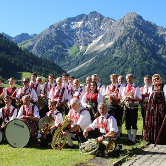 A group of musicians in traditional attire stands in front of a mountainous landscape. They are holding musical instruments and a flag in their hands. | © Musikverein d´Hirschegger | Jockl Metzger