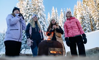 Eine Gruppe von Frauen steht um ein Feuer in einer verschneiten Landschaft. Im Hintergrund sind hohe, schneebedeckte Bäume und ein klarer blauer Himmel zu sehen.