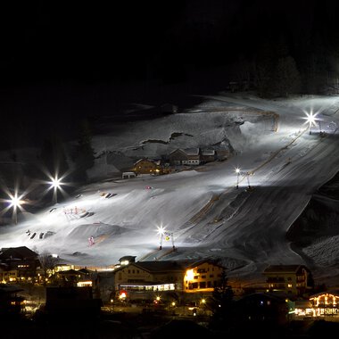 Ein wunderschön beleuchteter Skiort bei Nacht mit weißen Schneebergen. Die Lifte und Pisten sind gut sichtbar, während die umliegenden Gebäude sanft erleuchtet sind.