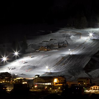 Ein wunderschön beleuchteter Skiort bei Nacht mit weißen Schneebergen. Die Lifte und Pisten sind gut sichtbar, während die umliegenden Gebäude sanft erleuchtet sind.