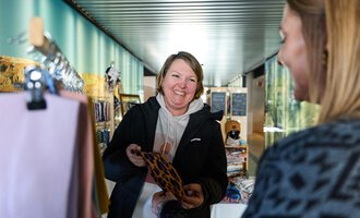 Eine lächelnde Frau zeigt ein Accessoire einer anderen Person. Im Hintergrund sind weitere Produkte in einem hellen, modernen Verkaufsraum zu sehen.