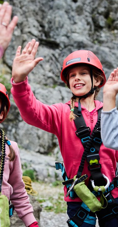 Three children in climbing gear greet an adult. They are wearing helmets and smiling against a rocky backdrop. | © Kleinwalsertal Tourismus | Oliver Farys