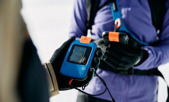 A person is holding a transceiver device in their hand. In the background, a snowy area is visible. | © Kleinwalsertal Tourismus | Oliver Farys