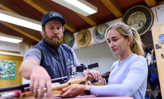 Ein Mann zeigt einer Frau, wie man ein Luftgewehr benutzt. Beide wirken konzentriert in einer sportlichen Umgebung. | © Kleinwalsertal Tourismus | Oliver Farys