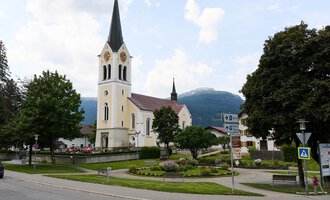 Eine malerische Kirche mit einem hohen Turm steht im Mittelpunkt eines kleinen Platzes. Im Hintergrund sind grüne Bäume und Berge zu sehen.