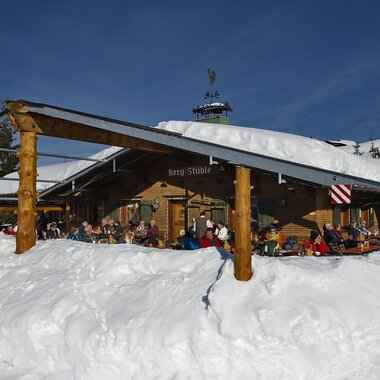 Eine gemütliche Berghütte im Schnee mit Gästen auf der Terrasse. Der Himmel ist klar und blau, und die Umgebung ist winterlich. | © Bergstüble | Michaela Beck Dornach