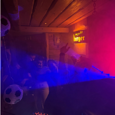 Eine lebhafte Party in einem gemütlichen Raum mit bunten Lichtern und Nebel. Menschen tanzen und scheinen viel Spaß zu haben.