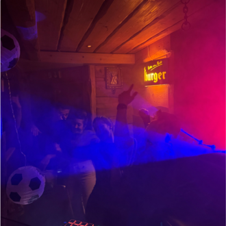 A lively party in a cozy room with colorful lights and fog.  
People are dancing and seem to be having a lot of fun.