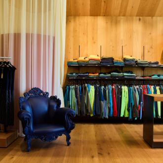 A modern store with a variety of clothing in vibrant colors. In the corner stands an elegant blue armchair.