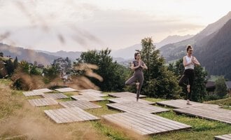 Zwei Frauen üben Yoga auf Holzplattformen in einer grünen Wiese. Im Hintergrund sind Berge und eine malerische Landschaft zu sehen.