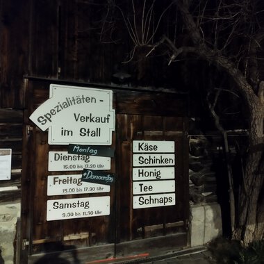 A wooden building with a sign for specialty sales in the barn. The signage indicates opening hours and offered products such as cheese and honey. | © Abler Spezialitätenverkauf im Stall | N. Lughammer