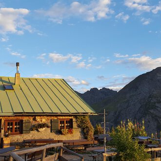 Eine gemütliche Berghütte mit grünem Dach in den Alpen. Im Hintergrund erstrecken sich majestätische Berge unter einem blauen Himmel. | © Alpe Kuhgehren | Lisa Hiesinger