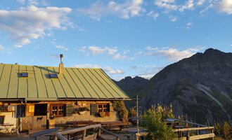 Eine gemütliche Berghütte mit grünem Dach in den Alpen. Im Hintergrund erstrecken sich majestätische Berge unter einem blauen Himmel. | © Alpe Kuhgehren | Lisa Hiesinger