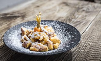 A black plate full of baked pieces, dusted with powdered sugar. A small piece of fruit adorns the presentation. | © Alpenhotel DAS KÜREN