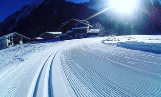 A snowy landscape with well-groomed cross-country ski trails. In the background, mountains and chalets can be seen, while the sun shines brightly.