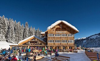 A cozy mountain restaurant surrounded by snow-covered trees. Many people are enjoying the winter atmosphere outside on the terrace. | © Auenhütte | Yorck Dertinger