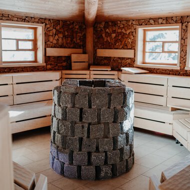 A cozy sauna room with wooden benches and large windows. In the center stands a stone oven. | © Verwöhn- & Wellnesshotel Walserhof| Aileen Melucci