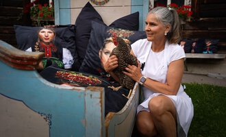 Eine Frau sitzt auf einer bunten Couch und hält ein Huhn in ihren Armen. Im Hintergrund sind künstlerisch gestaltete Kissen zu sehen. | © Annett Schwärzler Nails & Spa | Stefanie Haid