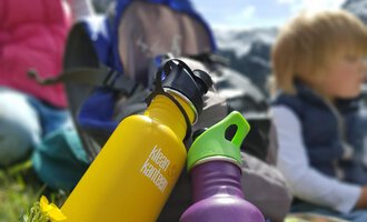 Zwei bunte Wasserflaschen stehen auf grünem Gras. Im Hintergrund sind Kinder und Berge zu erkennen. | © Austrian Ski- & Service Ranch | Trixi Leitner