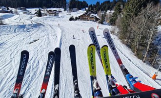A snow-covered ski slope with trees and cabins in the background. Skis are visible in the foreground. | © Austrian Ski- & Service Ranch | Trixi Leitner