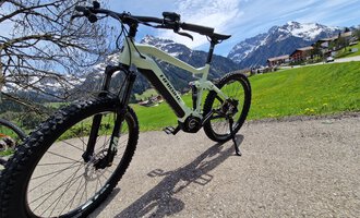 A modern mountain bike stands on a path in the midst of a picturesque mountain landscape. In the background, snow-covered mountains and green meadows can be seen. | © Austrian Ski- & Service Ranch | Trixi Leitner