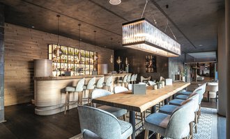 An elegant interior of a modern restaurant with a long wooden table and stylish chairs. In the background, there is a well-lit bar with a variety of drinks. | © A-ROSA Ifen Hotel Kleinwalsertal
