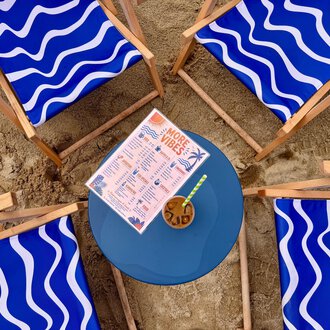 Four blue lounge chairs with wave design are set in the sand around a round table. On the table, there is a drink menu and a cocktail. | © MORE VIBES by Alex Lambsdorff