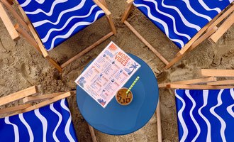 Four blue lounge chairs with wave design are set in the sand around a round table. On the table, there is a drink menu and a cocktail. | © MORE VIBES by Alex Lambsdorff