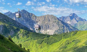 Eine Seilbahn schwebt über grüne Hügel und Berge. Der Himmel ist klar und blau, mit einigen Wolken. | © OBERSTDORF · KLEINWALSERTAL BERGBAHNEN