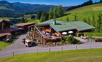 A picturesque mountain hut surrounded by green meadows and woods. On the terrace, people enjoy the sunny view of the mountains. | © Bergstüble | Jürgen Waffenschmidt