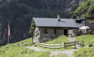Eine gemütliche Hütte in den Bergen, umgeben von grünen Wiesen. Im Vordergrund führt ein gepflasterter Weg zur Eingangstür, und eine Terrasse mit einem Sonnenschirm ist sichtbar. | © Bernhards Gemstelalp | Frank Drechsel
