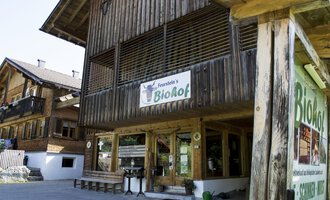 A wooden building with a sign that says "BioHof." Next to it are tables and chairs in the outdoor area, surrounded by other traditional buildings. | © Kleinwalsertal Tourismus