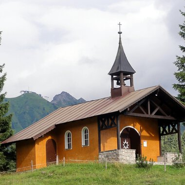 A small, orange church stands surrounded by green trees. In the background, mountains and a cloudy sky can be seen. | © Bruder Klaus Kapelle | Monika Gmeiner