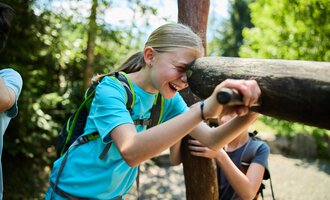 A cheerful child leaning against a wooden post, laughing, surrounded by a wooded area. The sun shines through the trees, creating a friendly atmosphere. | © Kleinwalsertal Tourismus | Oliver Farys