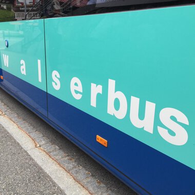 A bus with the inscription "walserbus" in a turquoise color scheme. The image shows the side of the bus on a road surface. | © Kleinwalsertal Tourismus