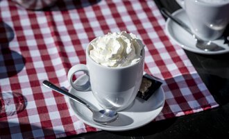 A cup with cream on a white saucer. The background is a red and white checked table runner. | © Kleinwalsertal Tourismus | Dominik Berchtold