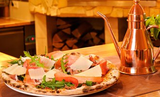 Eine frisch gebackene Pizza mit Schinken, Rucola und Parmesan. Neben der Pizza steht ein elegantes, kupfernes Ölbehältnis. | © Ristorante Trentino | Michael Monschau