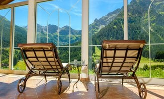 Zwei Liegestühle stehen vor großen Fenstern mit Blick auf grüne Berge. Der Raum wirkt einladend und hell. | © Hotel Leitner