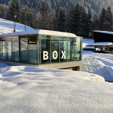 Ein modernes, gläsernes Gebäude mit der Aufschrift "BOX" steht inmitten einer schneebedeckten Landschaft. Im Hintergrund sind schneebedeckte Bäume und weitere Gebäude sichtbar. | © Sport Kessler GmbH