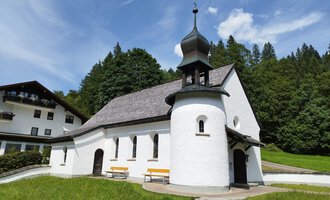 Eine kleine, weiße Kapelle mit einem schwarzen Dach inmitten von Grün. Im Hintergrund sind Bäume und eine Ferienunterkunft zu sehen. | © Kleinwalsertal Tourismus | N. Lughammer