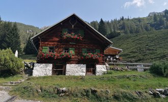 A rustic wooden house surrounded by green meadows and mountains. Flower boxes with colorful flowers adorn the windows. | © Fluchtalpe | N. Lughammer