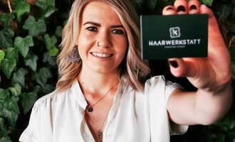 A smiling woman is holding a card for the hair workshop in her hand. In the background, green plants are visible. | © Friseur Haarwerkstatt | Franziska Diener