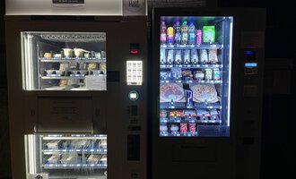 Two vending machines stand next to each other. The left machine offers frozen food, while the right one contains various drinks and snacks.