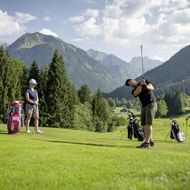 A group of golfers is practicing on a picturesque golf course with mountains in the background. The sun is shining, and the landscape is green and inviting. | © Golfclub Oberstdorf | ©kunst.joachimweiler.de