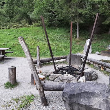 Ein gemütlicher Grillplatz im Freien mit einem großen Feuerkreis aus Steinen und Holz. Im Hintergrund steht eine Picknickbank auf einer grünen Wiese. | © Kleinwalsertal Tourismus | Veronika Senn