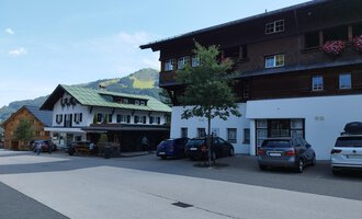 Ein ruhiges Dorf mit traditionellen Häusern und Autos auf der Straße. Im Hintergrund sind grüne Hügel sichtbar. | © Kleinwalsertal Tourismus | Nicole Lughammer