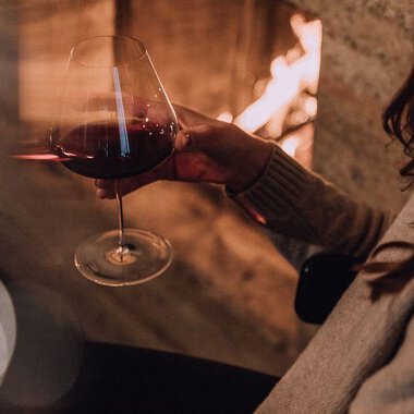 A person is holding a wine glass in their hand. In the background, a fireplace is burning, creating a cozy atmosphere. | © Naturhotel Chesa Valisa | Heldentheater